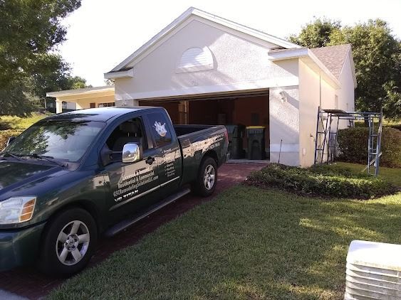 Green pickup truck parked in front of a cream-colored house with an open garage, on a sunny day