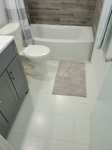 Bathroom with white tile floor, white tub, toilet, gray vanity, and neutral bath mat