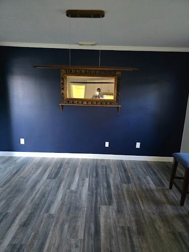 Dark blue wall with ornate mirror, gray wood-look floor, white trim, and a glimpse of a person in the reflection