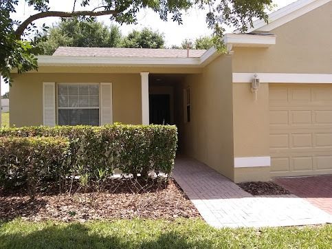 Tan house with brick walkway, entry under roof, hedge in front
