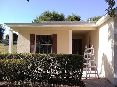 House exterior with ladder, front door, windows, bushes, and beige walls