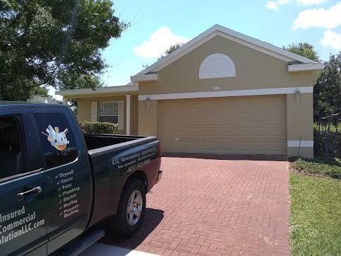 Green truck parked in front of a tan house with brick driveway on a sunny day