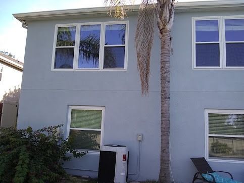 Back of a light blue stucco house with multiple windows, palm tree, and air conditioning unit