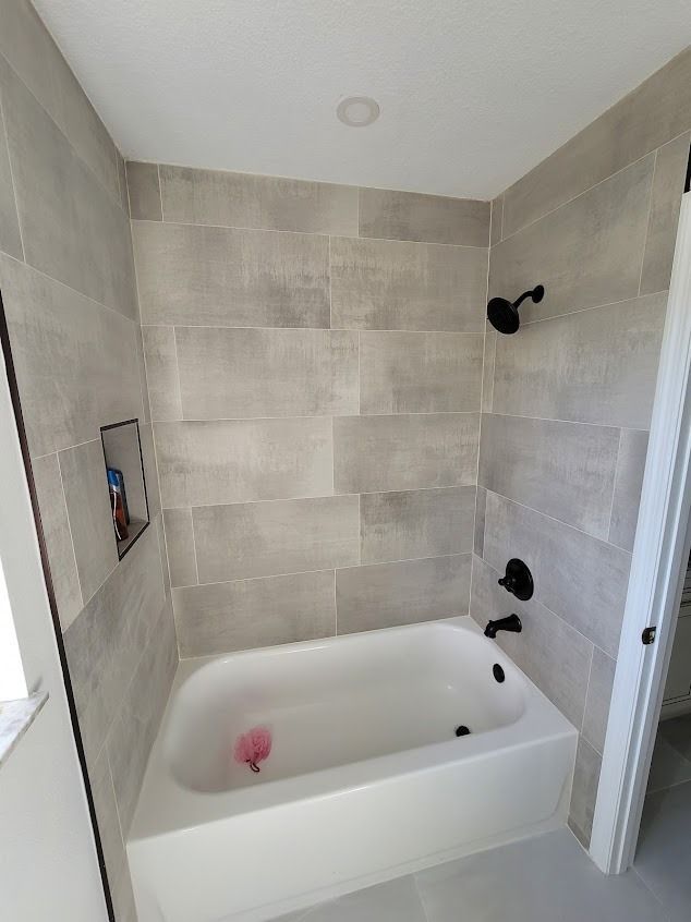 A bathroom with gray tile walls and white tub, black fixtures, and a built-in shelf