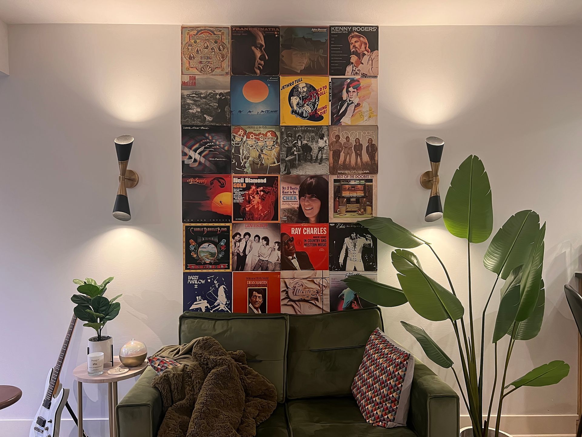 Cozy living room with green sofa, wall art collage, tall plants, and wall sconces.