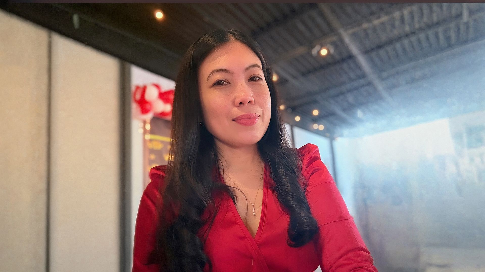 Woman in a red blouse sitting indoors, looking at the camera with a slight smile.