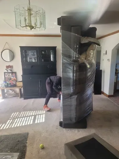Person wrapping large furniture in plastic wrap in a room with other furniture.