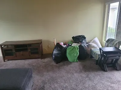 A room with a TV stand, bags of items, a chair, and a side table on a carpeted floor.