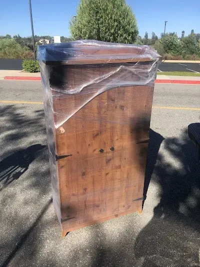 Wooden cabinet wrapped in plastic, outdoors. Brown wood, sunlight, near a curb.