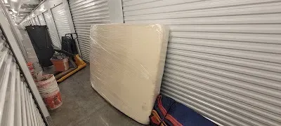 A mattress wrapped in plastic is leaning against a wall in a storage unit. Other items are visible in the background.