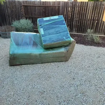A green-wrapped couch and box sit on gravel in a backyard, near a wooden fence and shrubs.