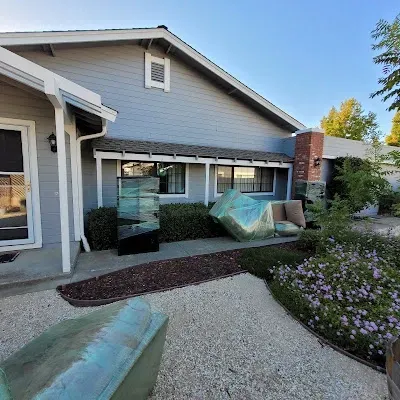 A house with a yard features outdoor artwork. One piece is a blue-green sculpture; another is a boxed chair.