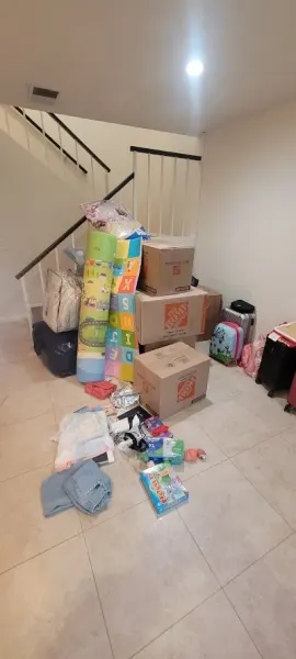 A cluttered room with boxes, bags, and children's items near a staircase. Beige tiled floor.