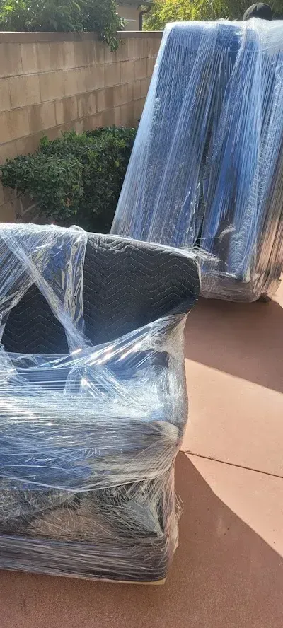 Wrapped furniture, likely for moving, sits on a patio with a brick wall and foliage in the background.