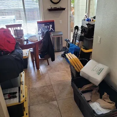 Messy kitchen with table, trash can, and boxes. Items like bags and sports equipment clutter the space.