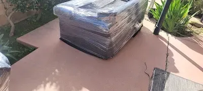 A large, plastic-wrapped object sits on a reddish-brown patio. Green vegetation and a pole are in the background.