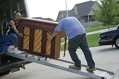 Two movers loading a piano into a moving truck, using a ramp on a sunny day.
