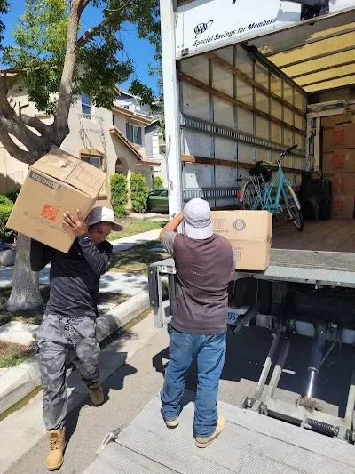 Two people moving boxes into a large truck on a sunny day.