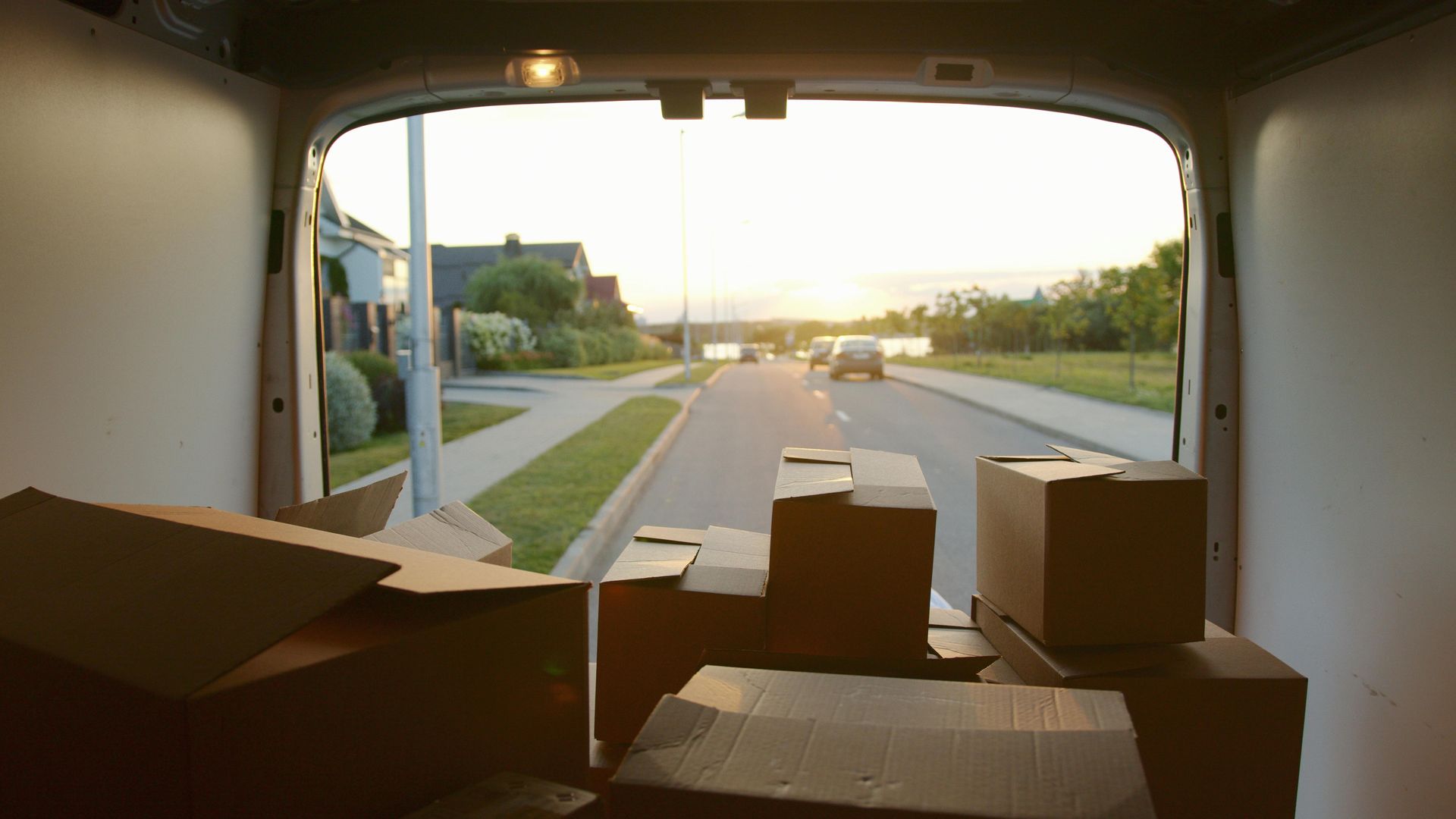 Interior view from back of a moving van filled with cardboard boxes, facing a street with houses.