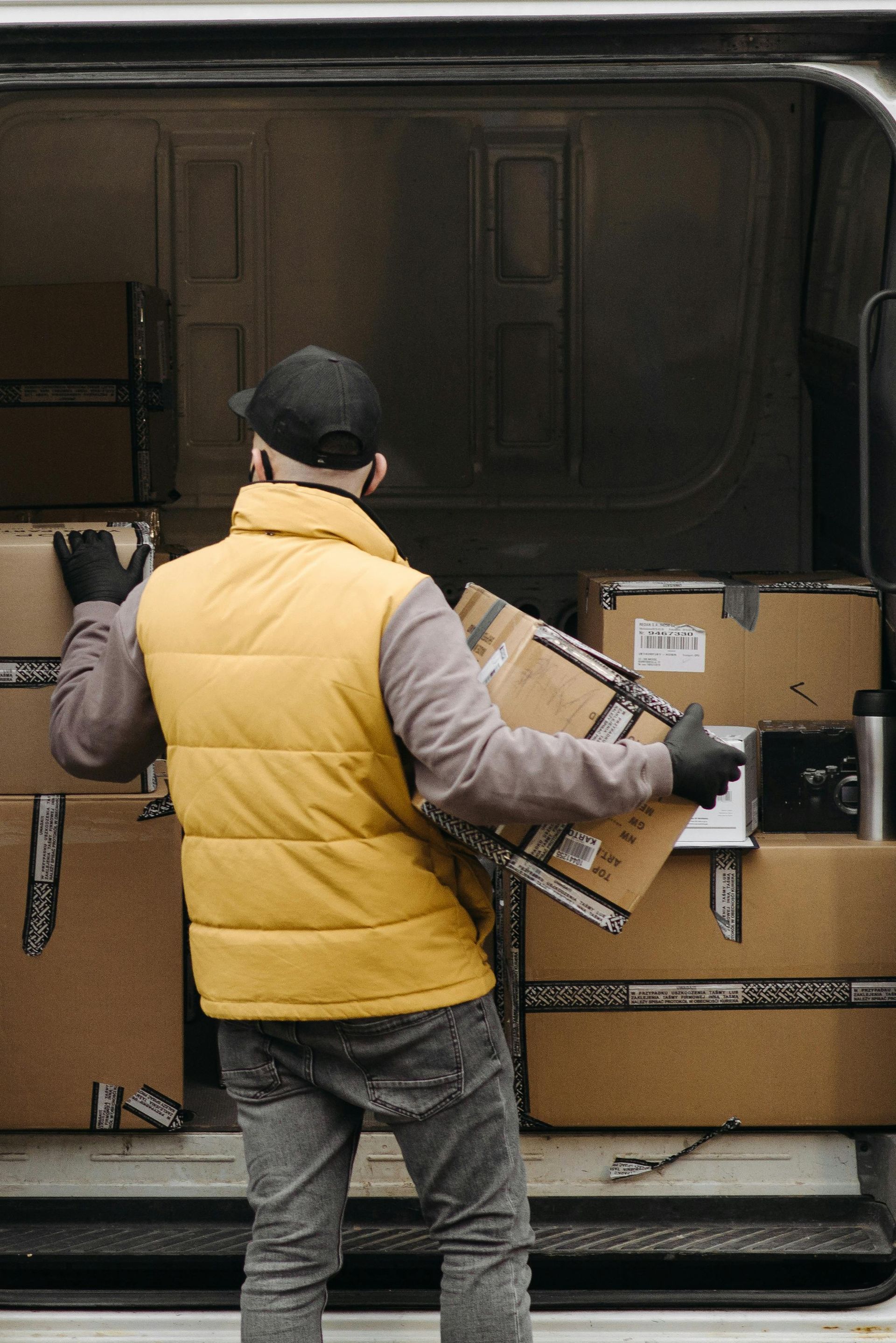 Delivery worker loading cardboard boxes into a van, wearing a yellow vest, black cap and gloves.