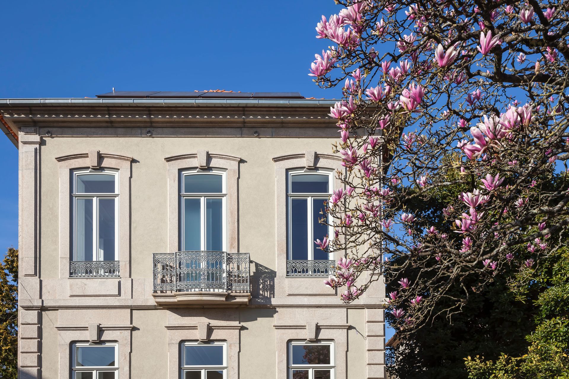 Facade of Casa Kala boutique guesthouse in Porto, Portugal, wrought-iron balconies, framed by blooming magnolia tree and garden. Designed by Januário Godinho, renovated by Ren Ito Arq.