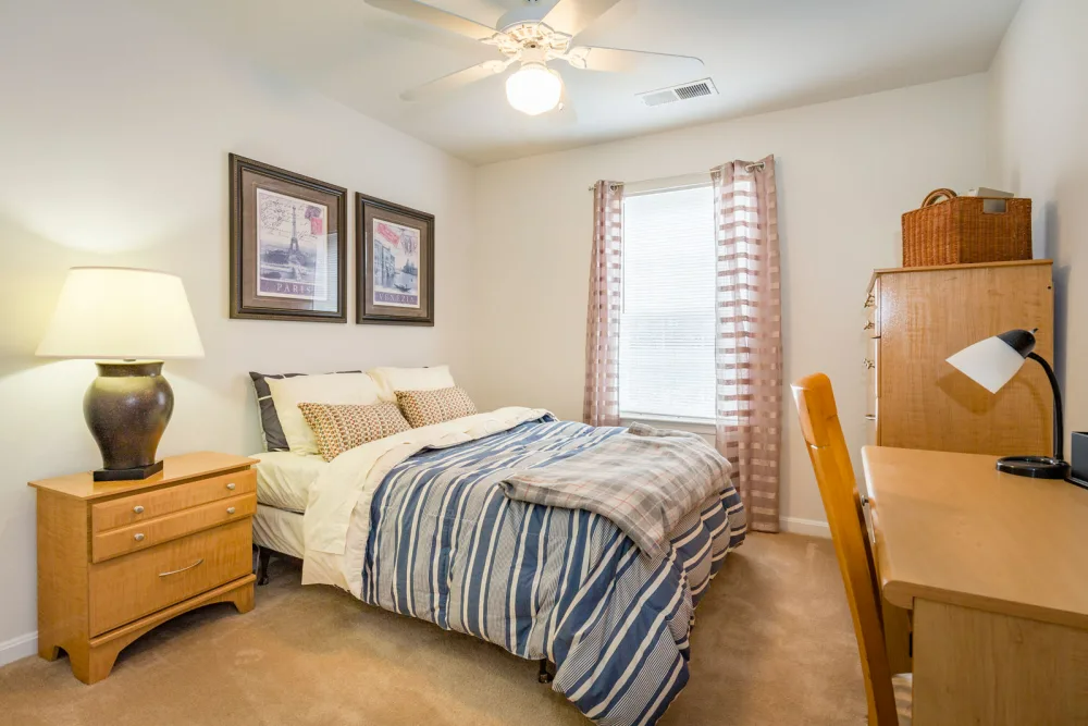 A bedroom with a bed, desk, nightstand, lamp, and ceiling fan.