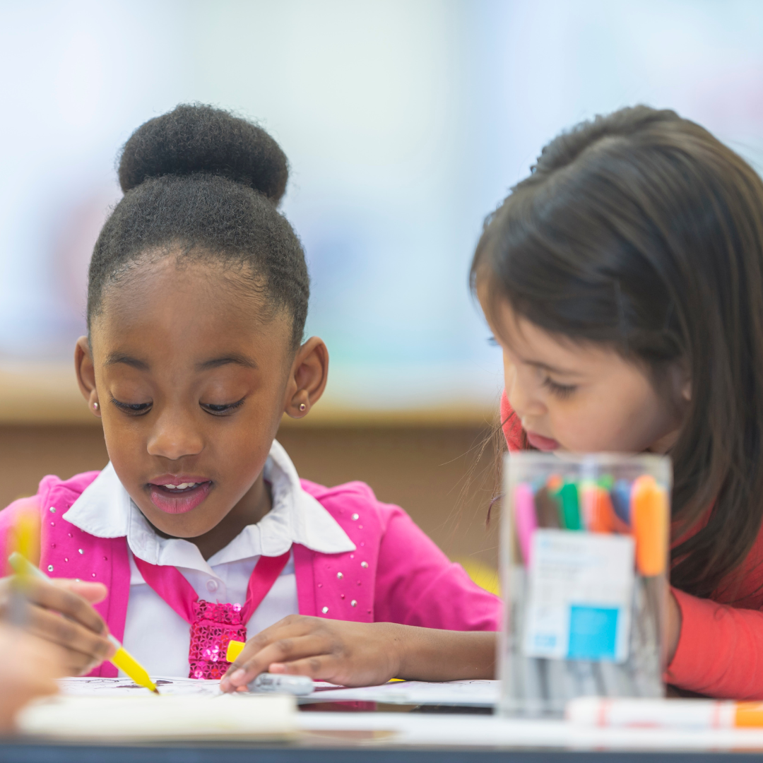 Two young girls are sitting at a table with markers in a classroom.