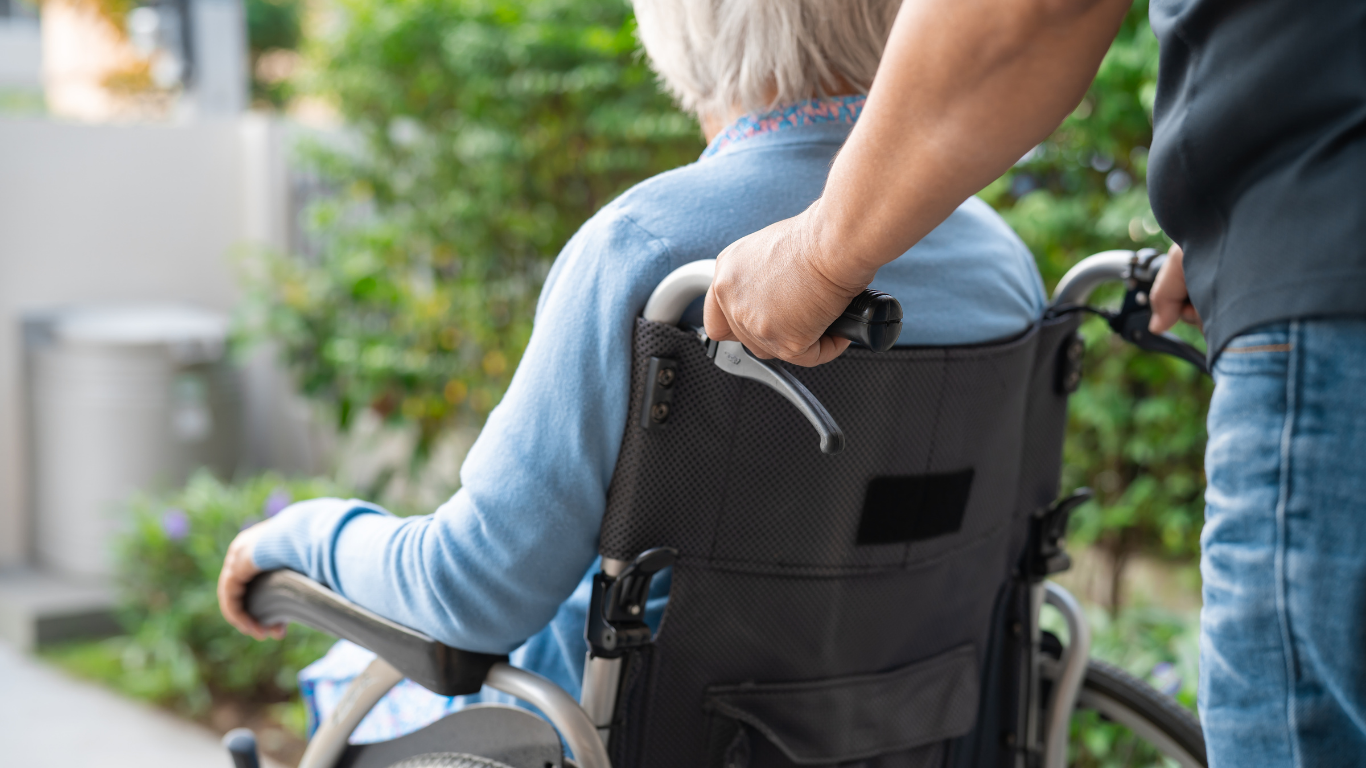 A person in a blue shirt sits in a wheelchair while being pushed by someone outdoors.