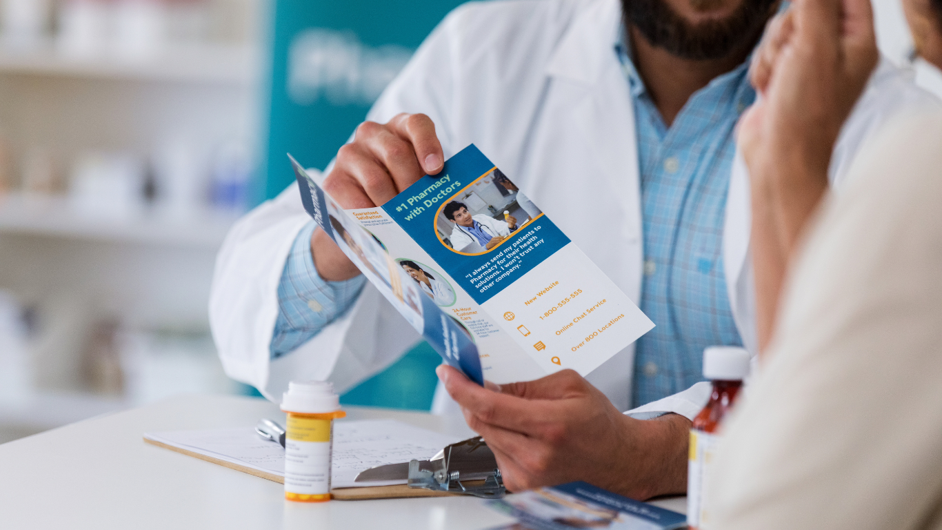 A pharmacist in a white coat shows a medical brochure to a patient across a pharmacy counter.