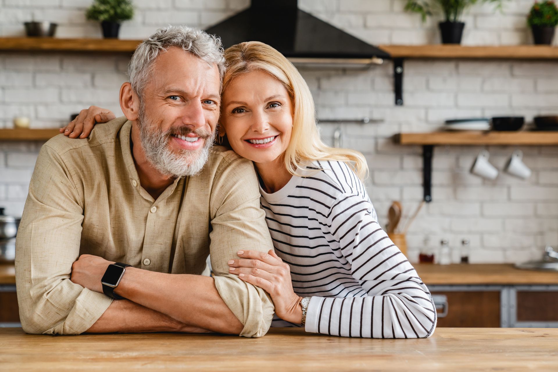 A smiling couple leans on a kitchen counter in a light-filled room with open shelves and brick walls.