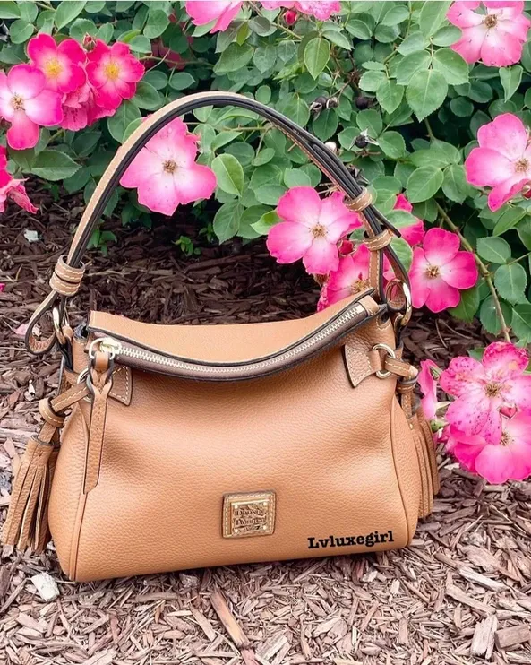 A brown purse is sitting in front of a bush of pink flowers.