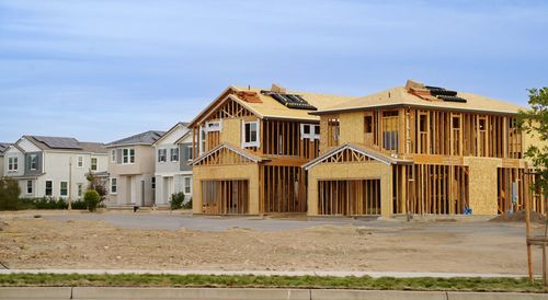 New houses under construction on a dirt lot; completed houses in the background. Blue sky.