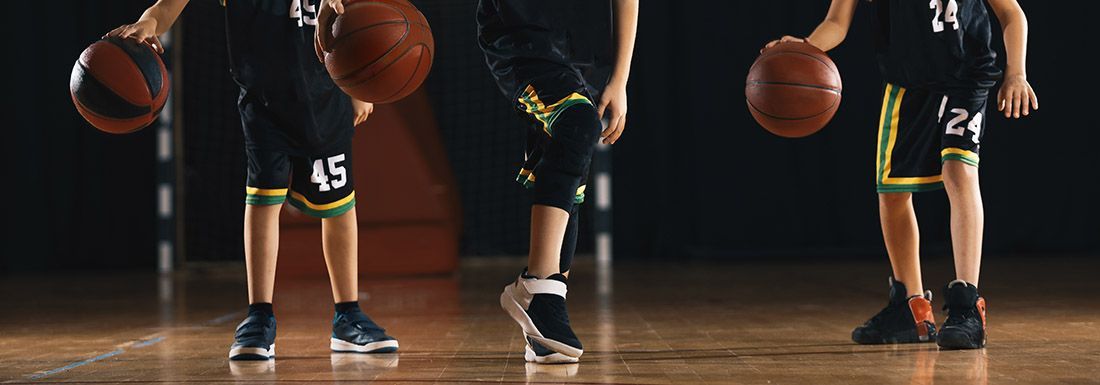 Torso, legs and feet of 3 basketball players, all bouncing balls. Each is wearing a different type of sneaker.