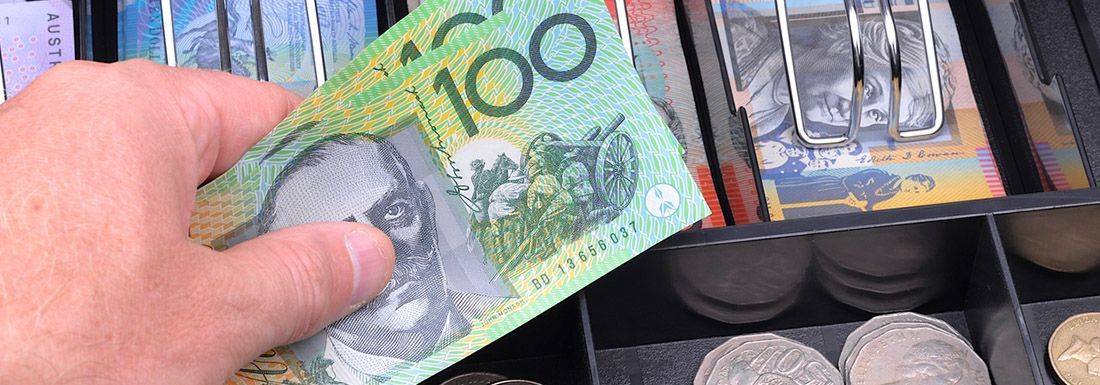 A close up of an open cash register drawer with Australian currency and a hand holding $100 notes as if to be taking a payment from a customer.