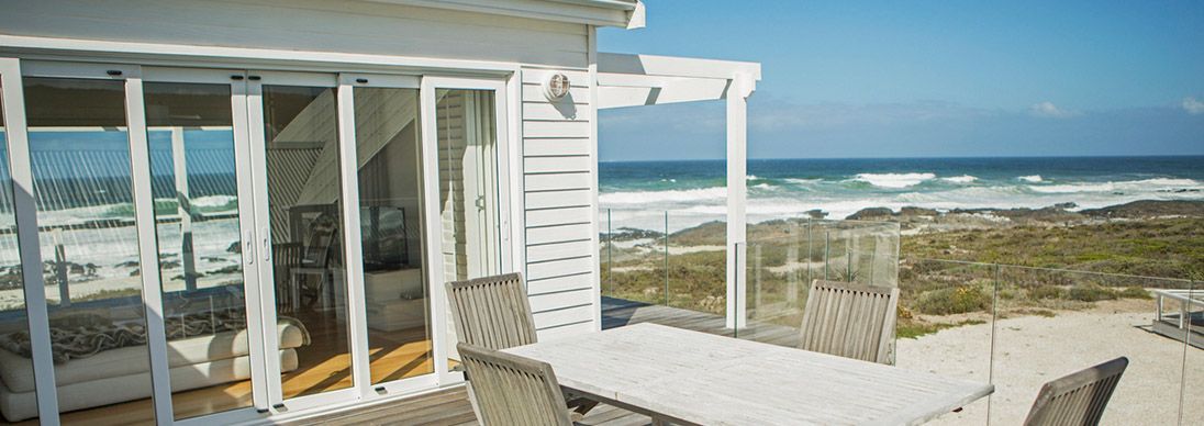 A holiday home with views over the ocean from the outside. There is a deck with a table and chairs.