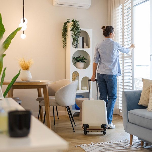 A woman with a rolling suitcase in a holiday home, opening blinds.