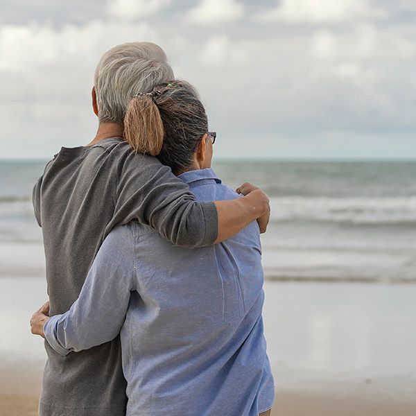 An older couple on the beach with their arms around each other, facing the water.