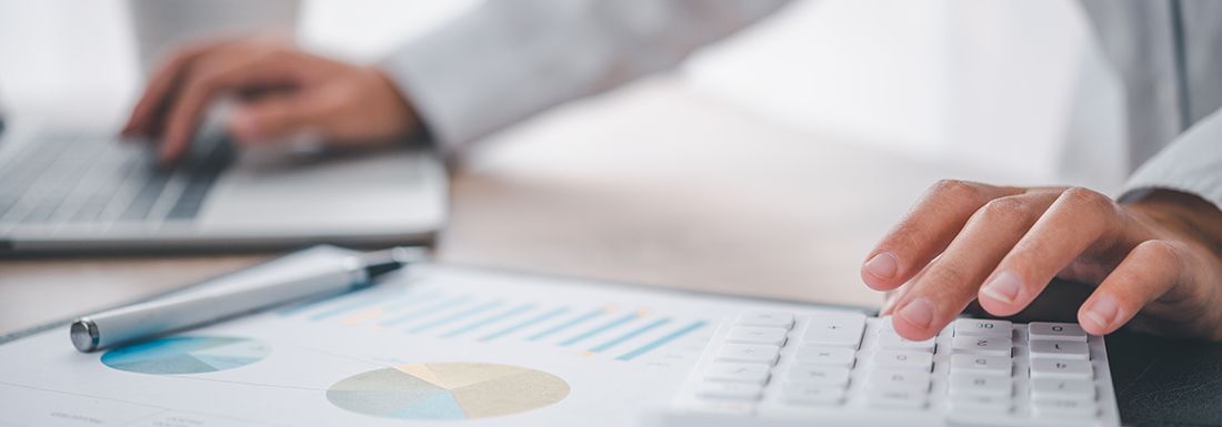Close up of a man's hands working with a laptop and calculator. There is a print out of pie charts in front of him.