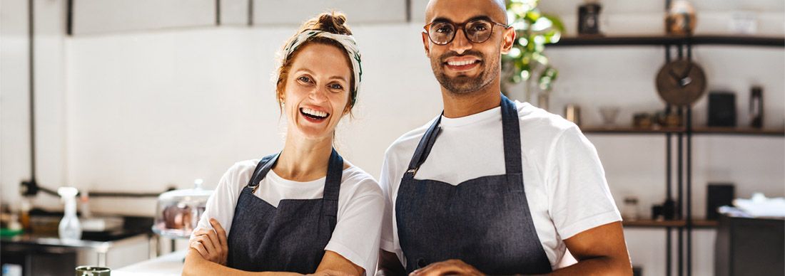 A women and a man cafe owners wearing white t-shirts and blue aprons. They are smiling and looking at the camera.