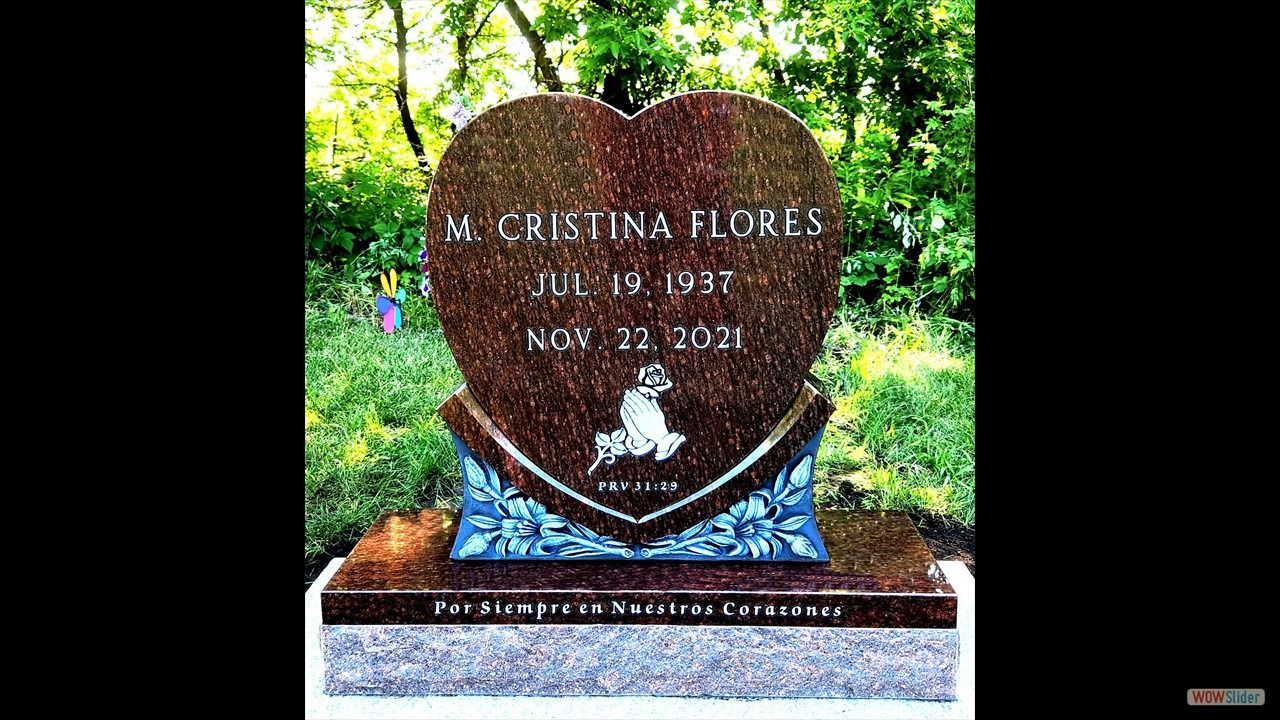Heart-shaped headstone for V. Cristina Flores, dates 1987-2021, with flowers and a small angel in a cemetery.