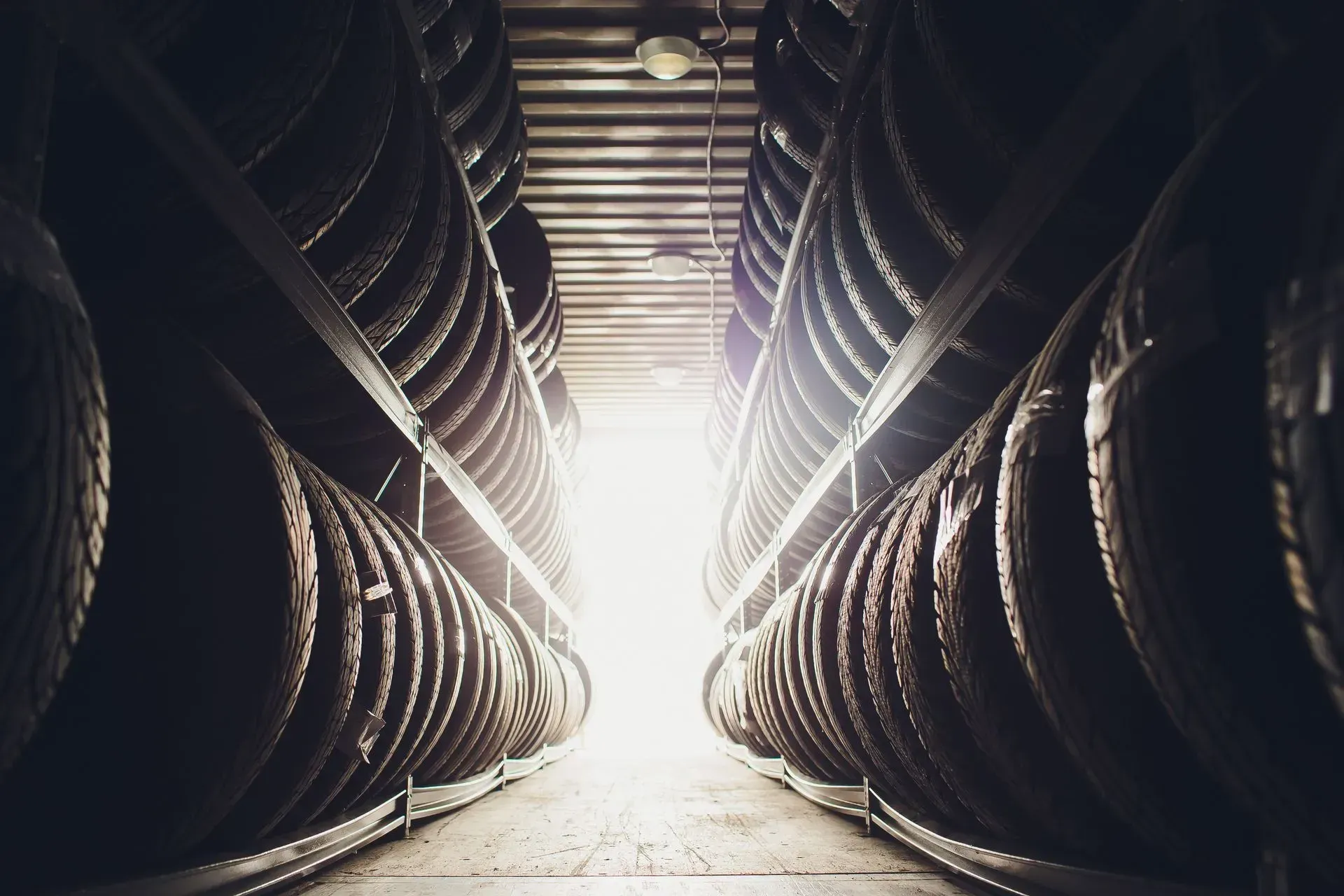 Due file di pneumatici impilati fiancheggiano uno stretto corridoio scarsamente illuminato di un magazzino, che conduce a una luce intensa in fondo al corridoio.