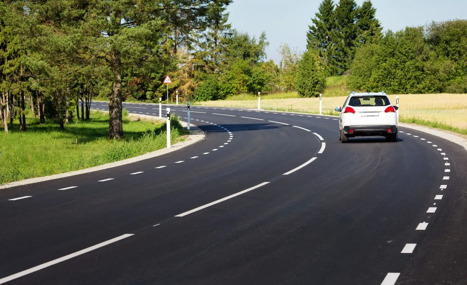 In una giornata di sole, un'auto argentata percorre una strada asfaltata curvilinea, fiancheggiata da alberi verdi e campi.