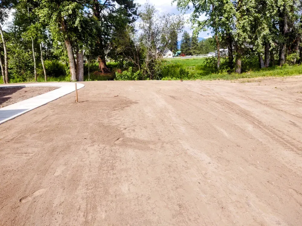 A cleared dirt lot with a concrete sidewalk, trees, and a distant grassy hill.