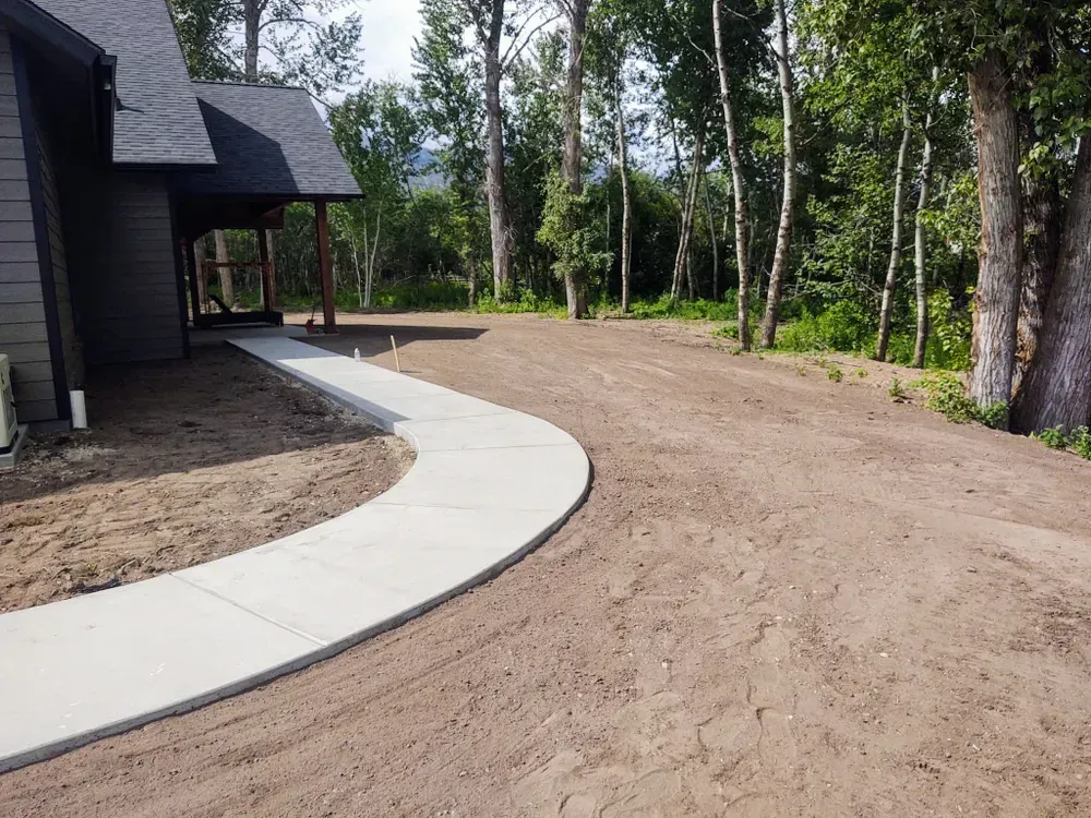 Concrete walkway curves through dirt yard beside a house and trees.