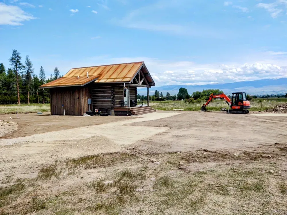Log cabin under construction; excavator nearby, blue sky, gravel pathway.