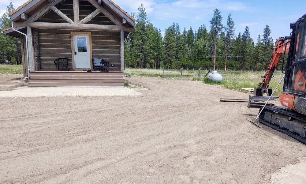 Gravel driveway being graded near a log cabin with an excavator; tall trees and sky in the background.