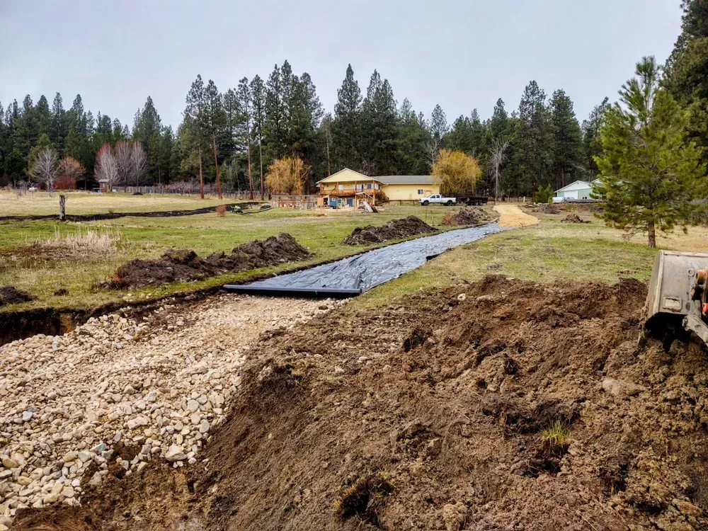 Construction site: a trench with gravel, black fabric, and dirt in a field with trees and a building in the background.