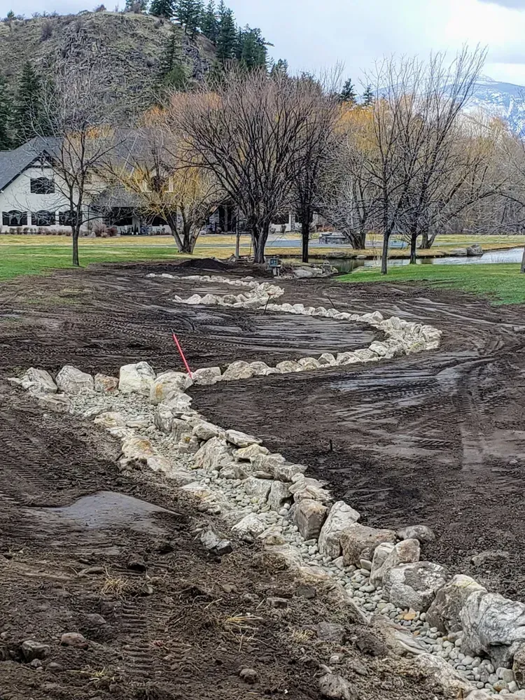 A meandering rock-lined ditch on a muddy lawn; bare trees, a house, and a distant mountain.