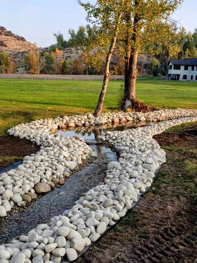 Stream lined with rocks flowing through a grassy area with a tree and hillside in the background.