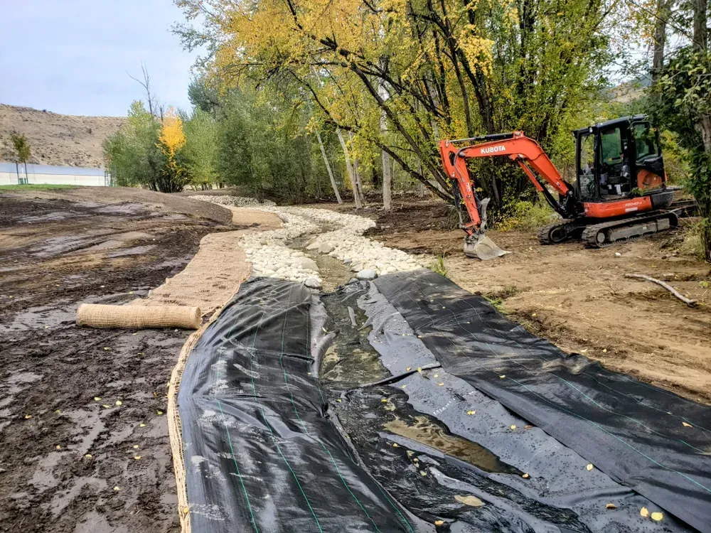 Construction of a creek bed with rocks and black fabric, using an orange excavator. Trees in background.